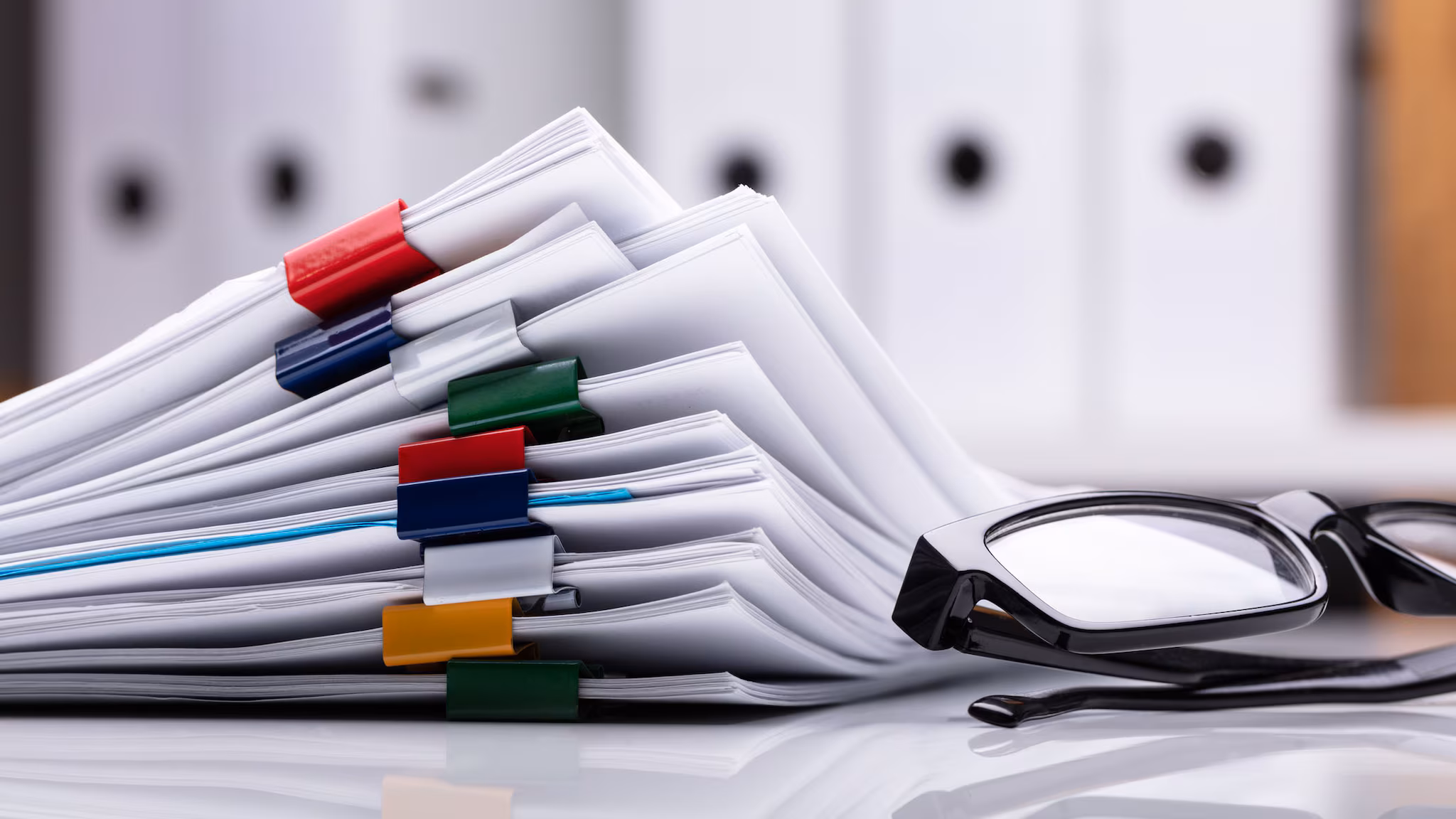 Stacked papers on desk next to reading glasses.