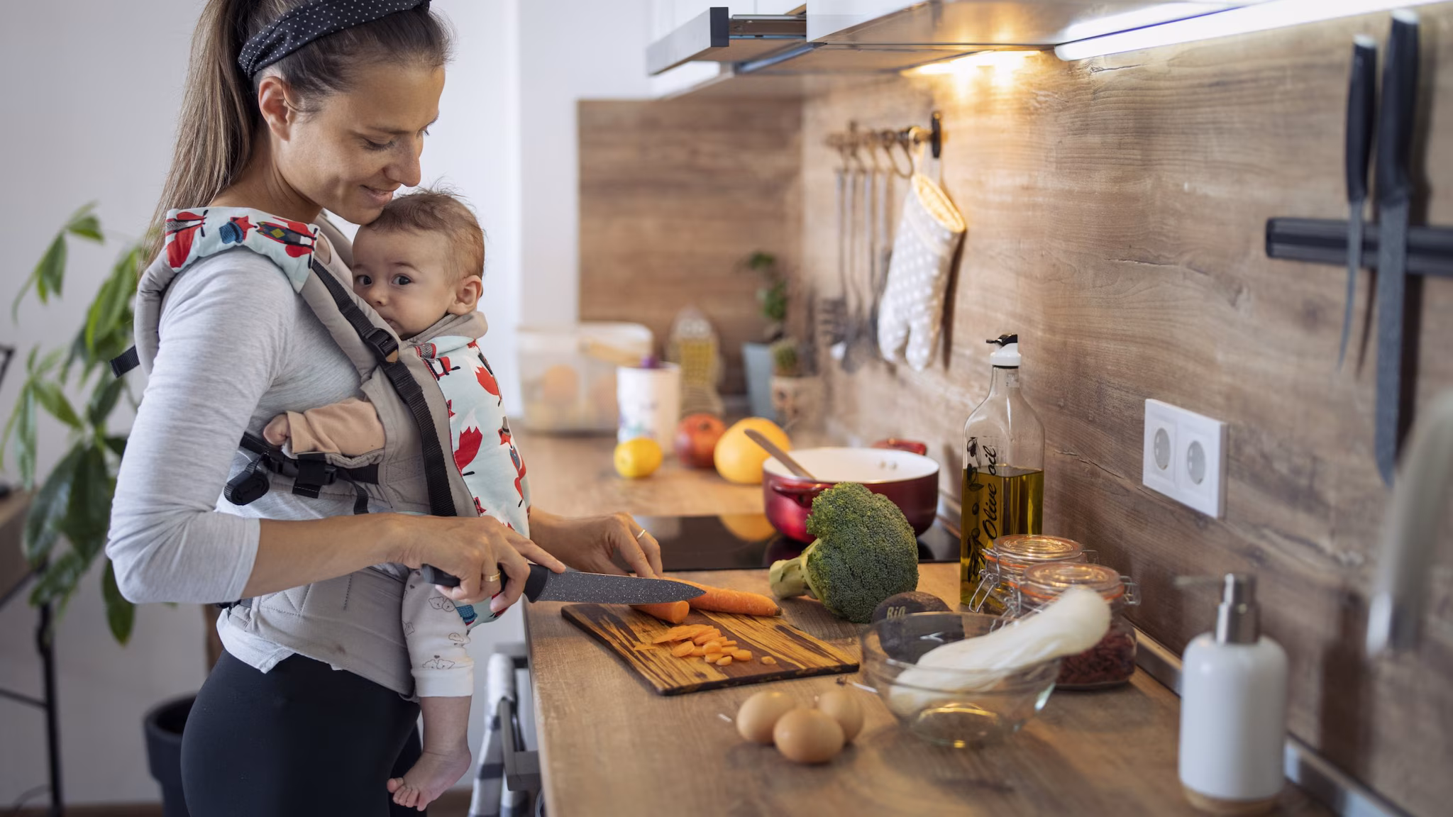 Mother cutting carrots in kitchen while a baby is in a carrier on her chest