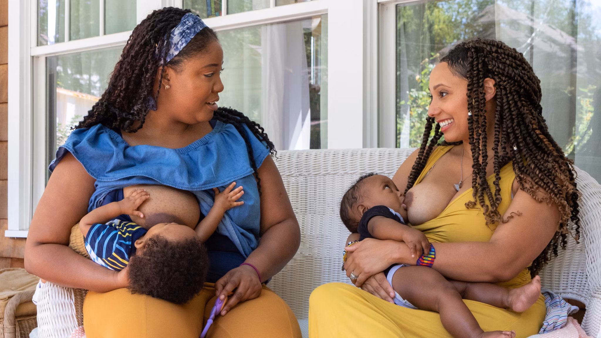 Two women breastfeed two children while sitting on a porch outside a house.