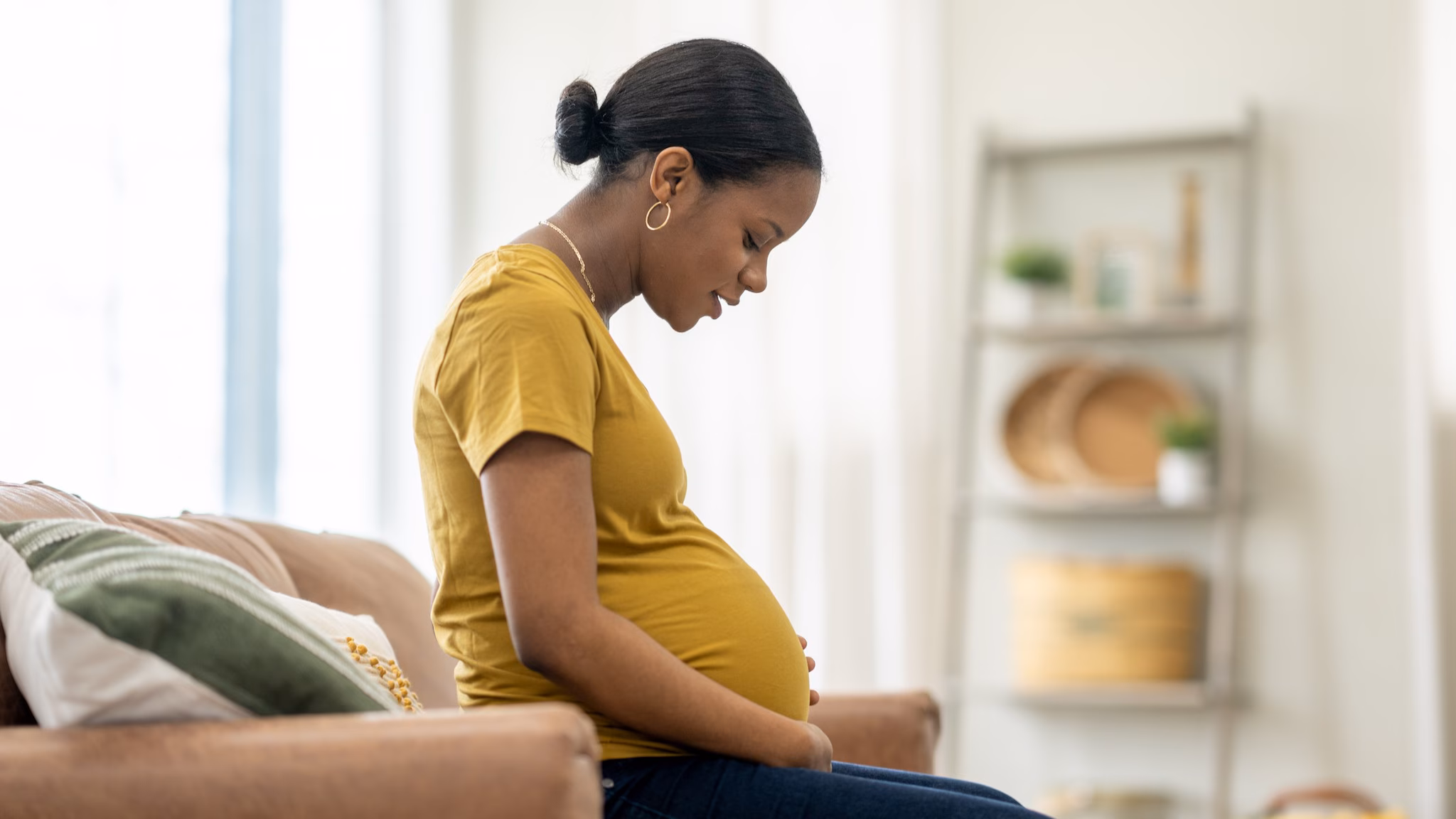 Pregnant Pregnant person in a yellow shirt sitting on a couch looking at their stomach.