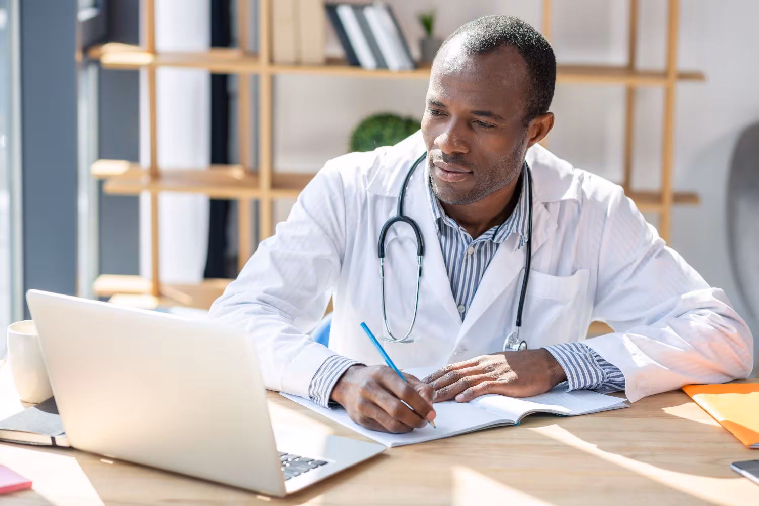 A doctor looking at his computer while taking notes at his desk.