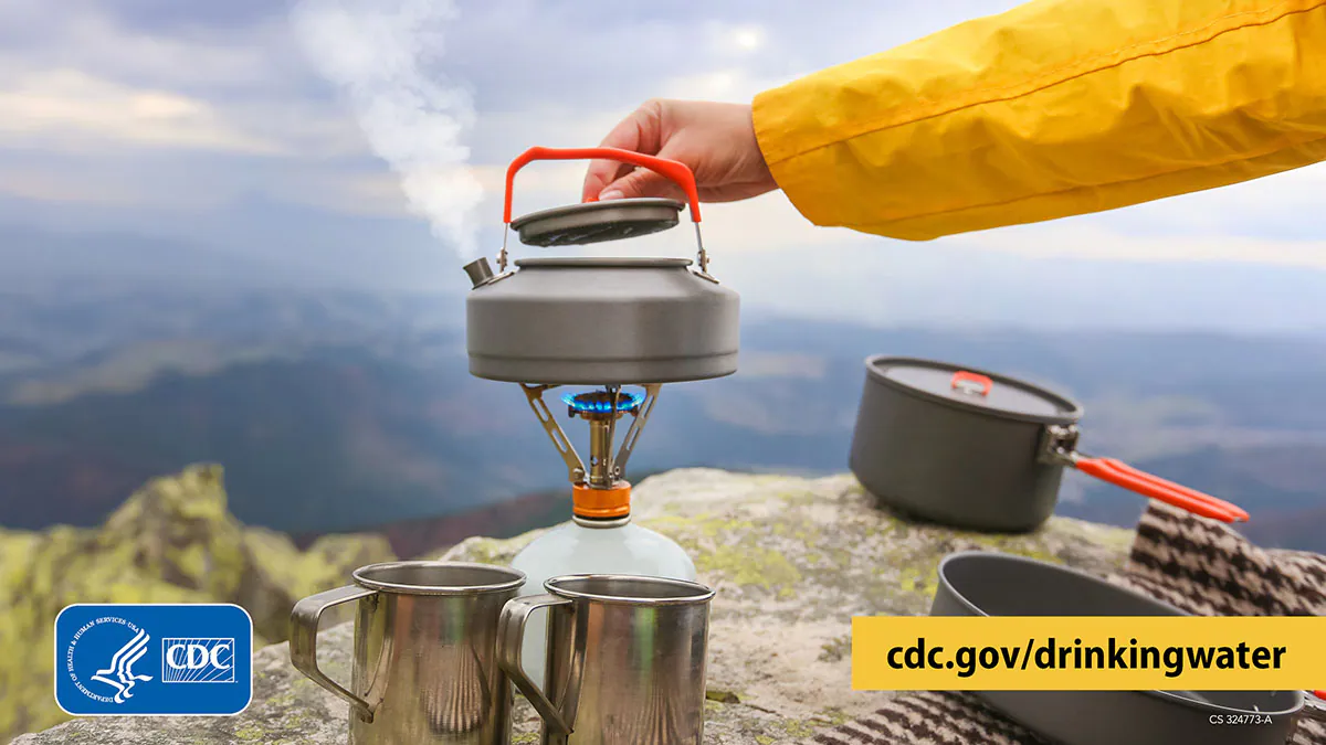 Boiling water on a mountain 1200x675 Hiker on a peak overlooking mountain ranges. Hiker lifts the lid of a kettle of water boiling on a portable stove, with pots and mugs nearby.