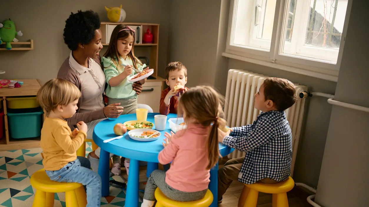 Teacher_children_eating Teacher and young children sitting at a table eating nutritious foods.