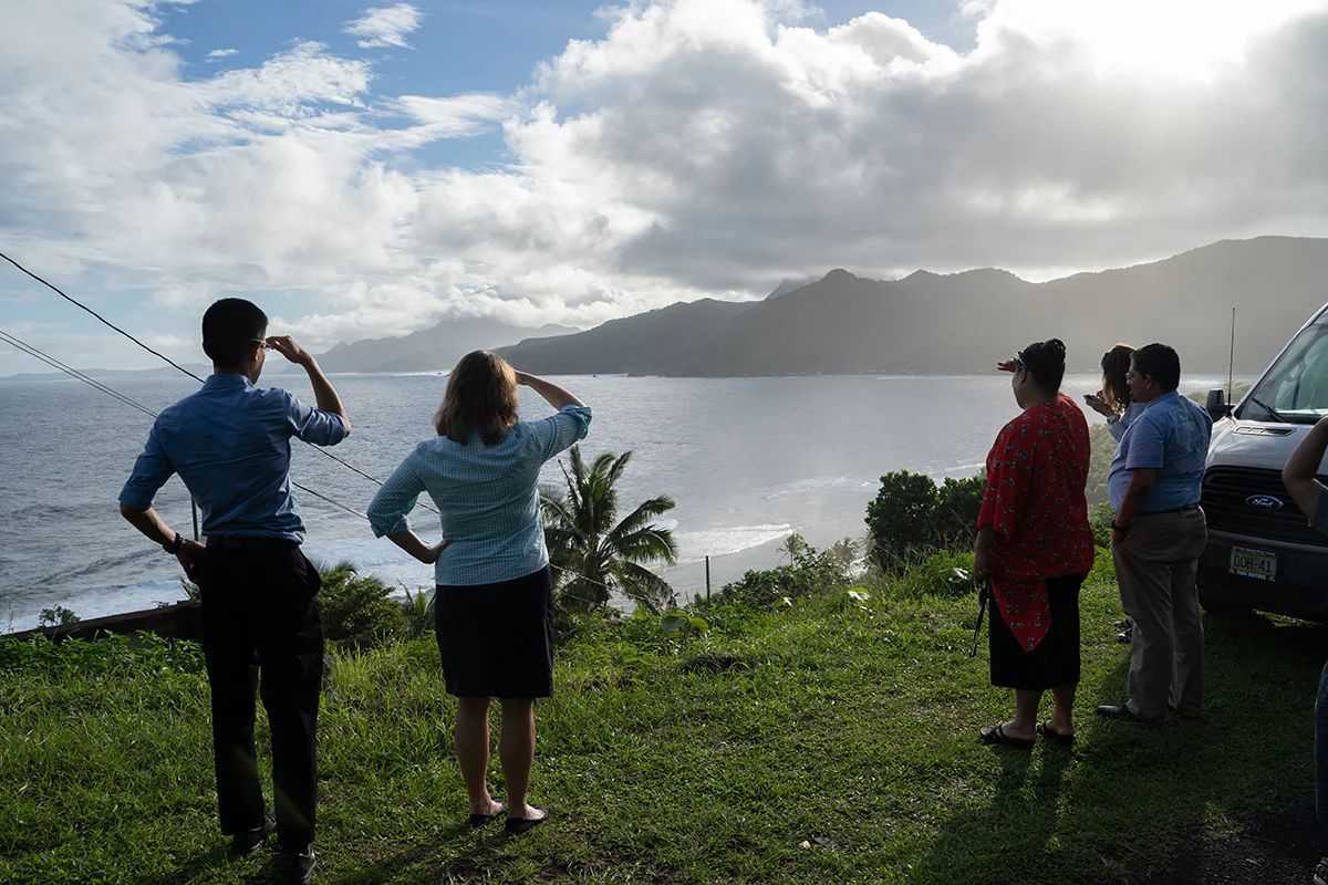 howard-chiou-american-samoa 5 people stand with their backs to the camera, facing an ocean overlook with palm trees. Two of the people salute toward the view.