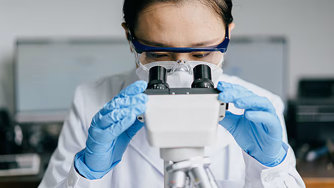 A person wearing a lab coat, safety goggles, a face mask, and blue latex gloves. A person wearing a lab coat, safety goggles, a face mask, and blue latex gloves.