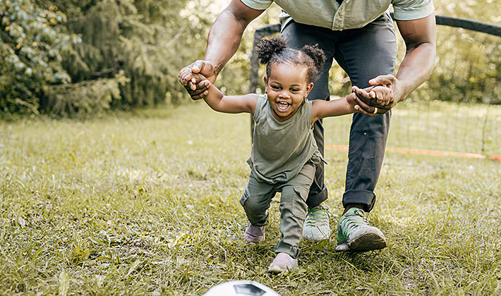 Caregiver helping a toddler run towards a soccer ball. Caregiver helping a toddler run towards a soccer ball.