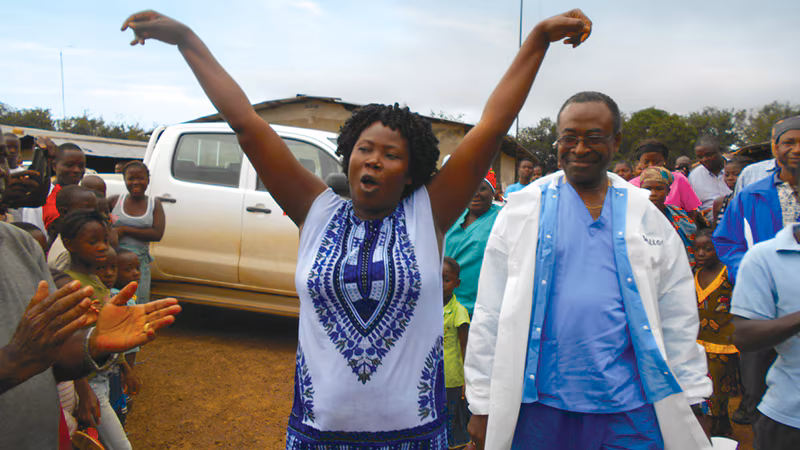 A joyful woman with arms raised celebrates with a doctor and community members in the background.