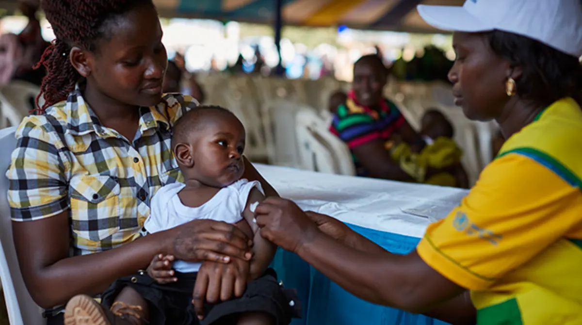 Malaria Vaccine RTS, S MVIP A woman in a yellow and green uniform administers a malaria vaccine to a baby held by his mother.