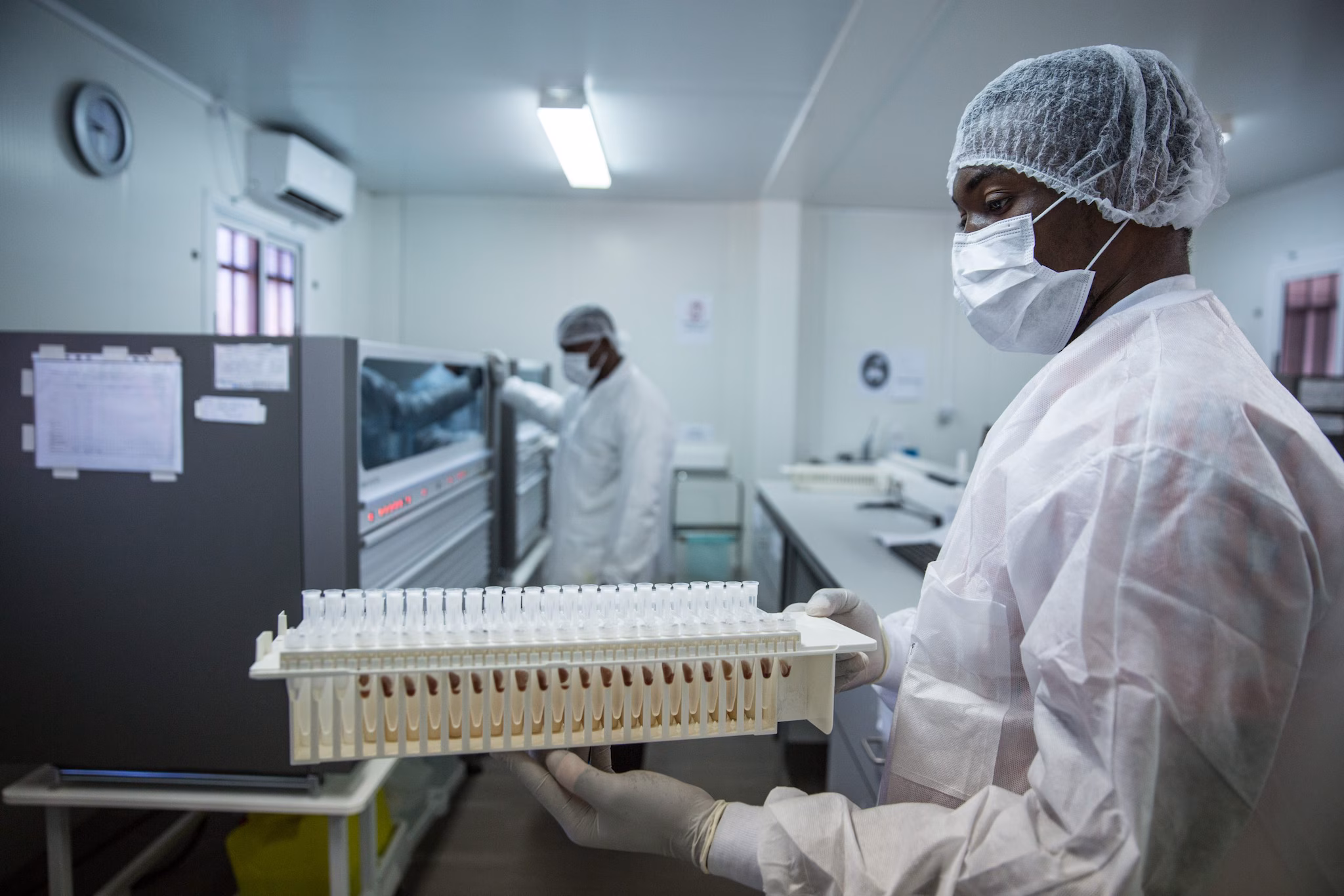 A man wearing white lab gear, blue gloves, a mask, and hair protection stands in a laboratory.