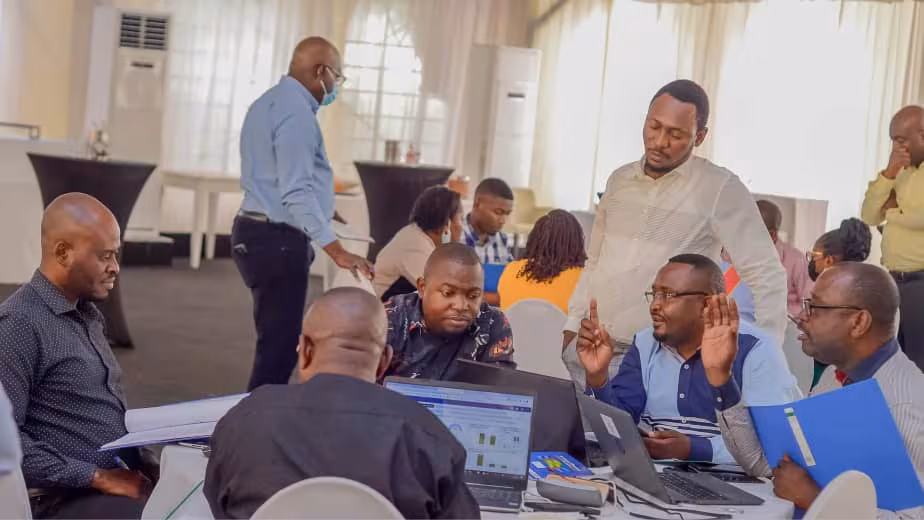 DRC_HIVTraining_FieldPhoto_2021 A room with many people sitting at round tables, talking with one another, reading, and using laptops.