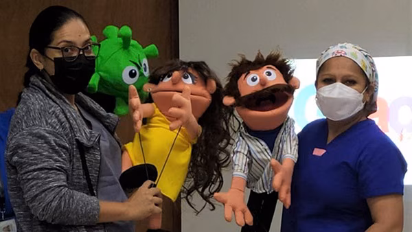 Two masked health workers pose with hand puppets. The puppets resemble a coronovirus with eyes and a mouth, a child, and a man.