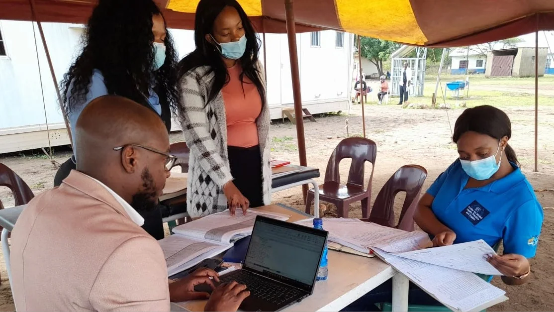 Eswatini_SIMSvisit_Photo Four people stand and sit around a table under an outdoor tent.