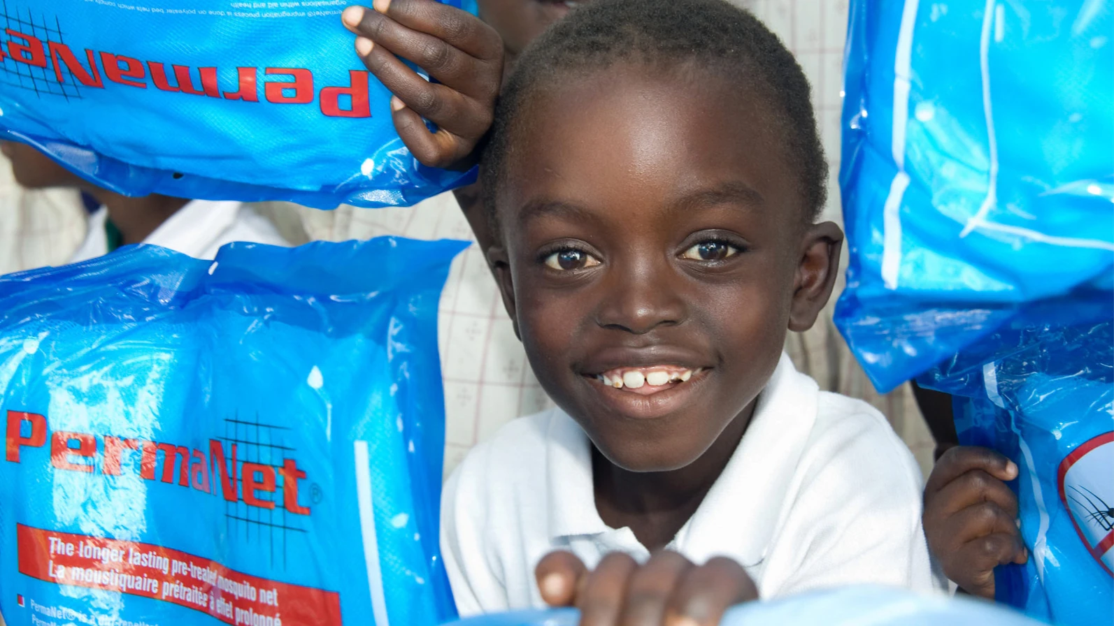 Kenya-child-bednets-16x9 Photo of a young child smiling while people hold up packages of wrapped bed nets around him.