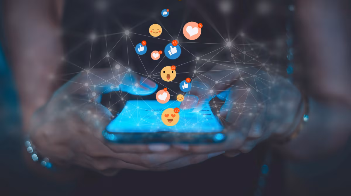 Close up of a person using smartphone with social media emojis floating above the phone screen.