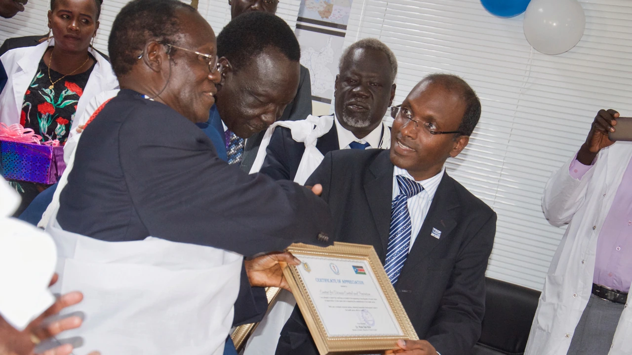 SouthSudan_AwardToSudhirBunga_Photo_2019 Two people hold a framed certificate of appreciation. Other people look on nearby.