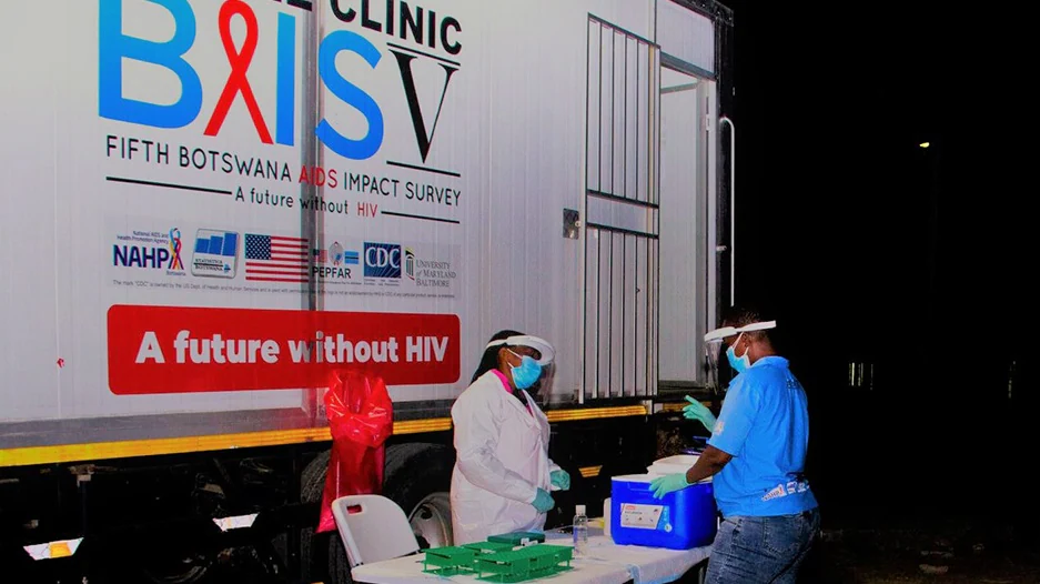 — title missing — Three individuals in personal protective equipment stand in front of a BAIS V truck with coolers of medical samples.