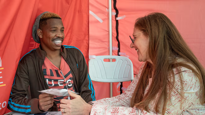 Archibald, 29, wearing a red shirt and oversized hat, sits beneath a giant tent waiting to take a test that could change his life