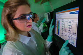 NGS_Image_Revised343x227 A woman with short blonde hair wears safety goggles and a lab coat while looking at lab results on a screen