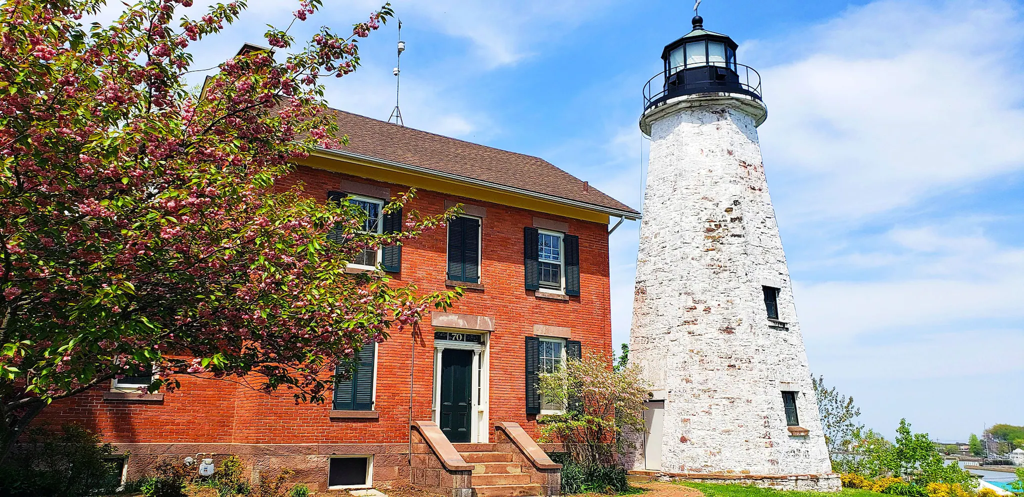 A red brick building with black ss next to an old white lighthouse with a black top, surrounded by blooming trees and a clear blue sky.