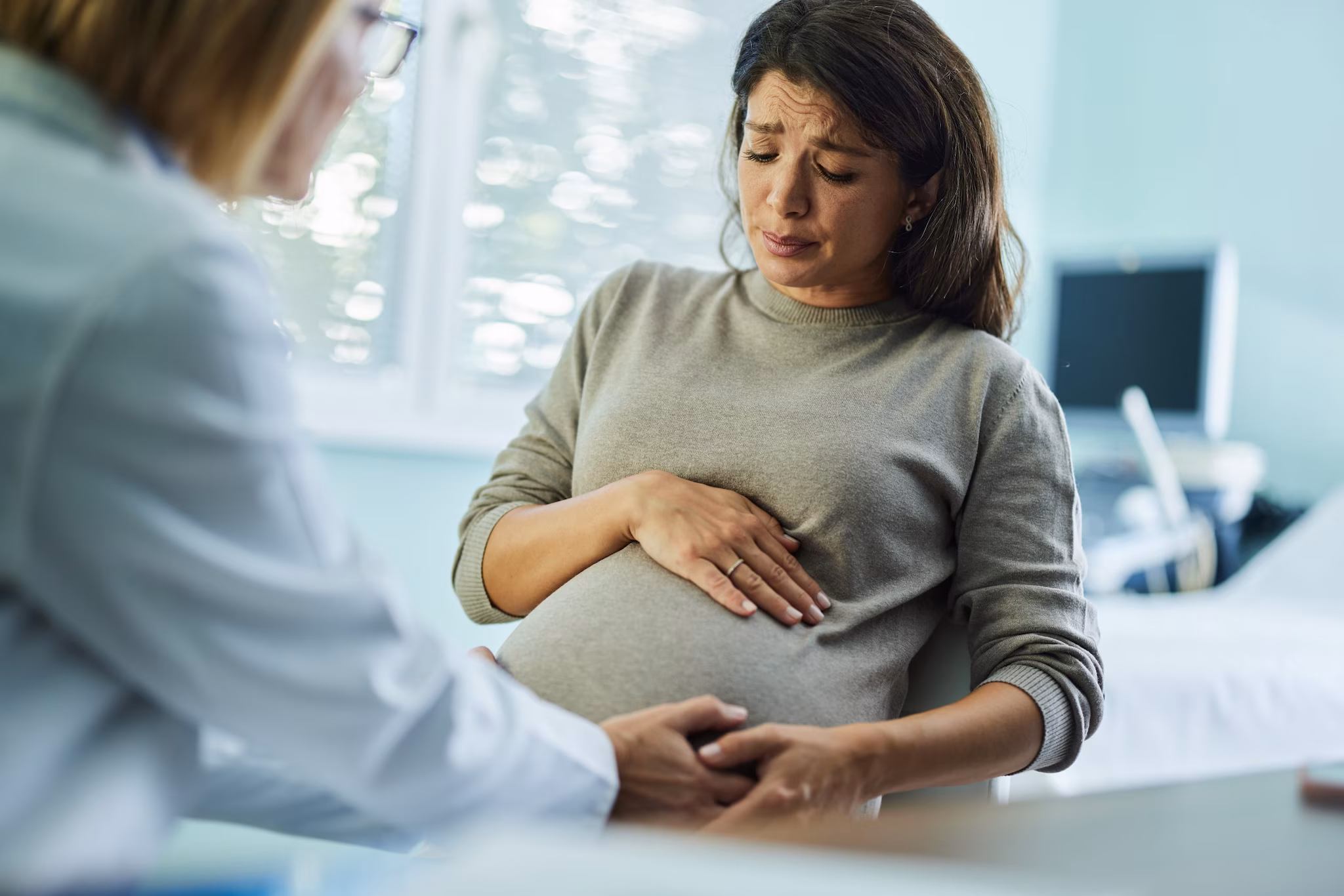 Patient care A pregnant person holding their pregnant stomach and looking worried while speaking to a doctor in a medical office.