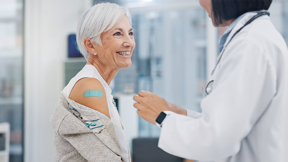 Older woman with white hair showing a teal bandage on her arm and smiling at a doctor wearing in white coat