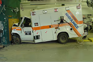 This picture is set inside an industrial looking space with concrete floor and wires and equipment attached to the walls. In the background you can see a pegboard wall with tool neatly arranged on it. In the foreground is an ambulance which has crashed head-first into a wall at the spot where there is a reddish brown plate just a little bit wider and taller than the grill of the ambulance.