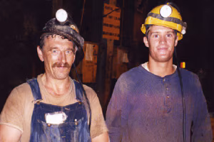 Two men look directly at the camera and grin. They are both very dirty. One man in overalls and t-shirt wearing a black miner’s hardhat with a light attached to the front looks to be about 50. Standing very close to this man is a younger man, probably in his twenties. He is wearing a dirty blue shirt and a yellow miner’s hard hat with a light attached to the front and a smaller one just above his right ear.