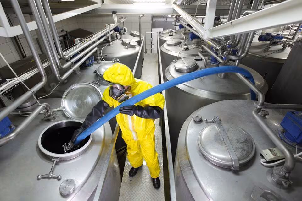 Chemical Manufacturing Worker Worker in full PPE in a chemical factory places a hose in the top of a vat.