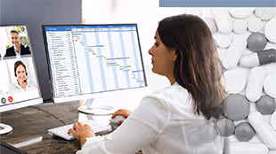 Woman in white shirt sitting at desk looks at a video conference call on one monitor and a spreadsheet on another monitor.