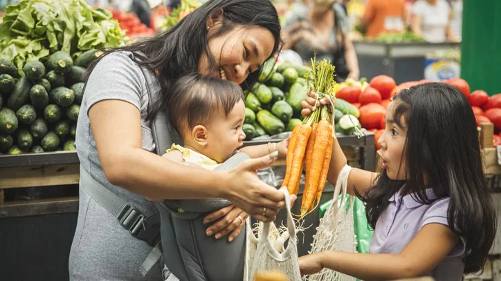— title missing — Mother with two young children at a produce store. One child is putting a bunch of carrots in a shopping bag.