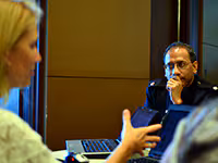 A man listens to a woman speaking at a table filled with laptops.
