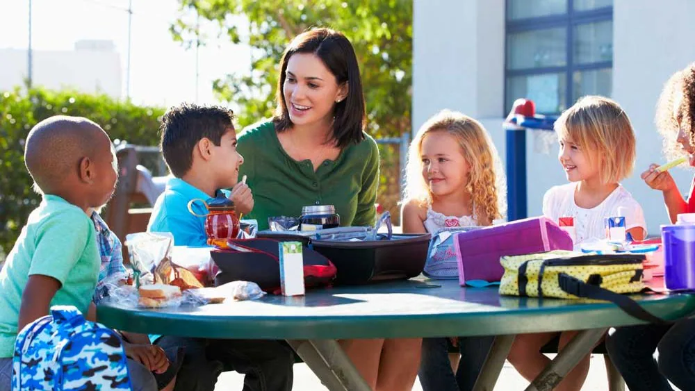 — title missing — Adult and five children at table eating a meal outside.