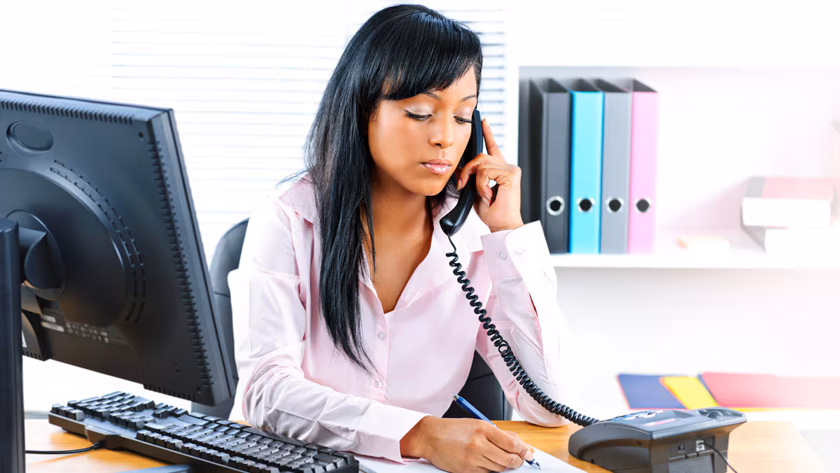 Woman on telephone taking notes A woman seated at a desk listens on a telephone while taking notes.