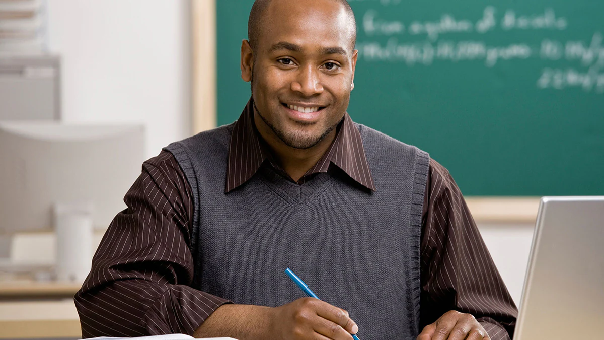 Teacher Sitting at Desk with Pen in Hand A teacher sits on a desk, holding a notebook in his hands.