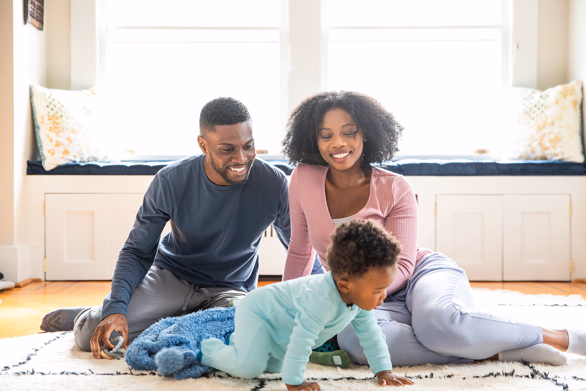 Parent watch baby crawl Two smiling parents sitting on the floor together in home watching their baby crawl.