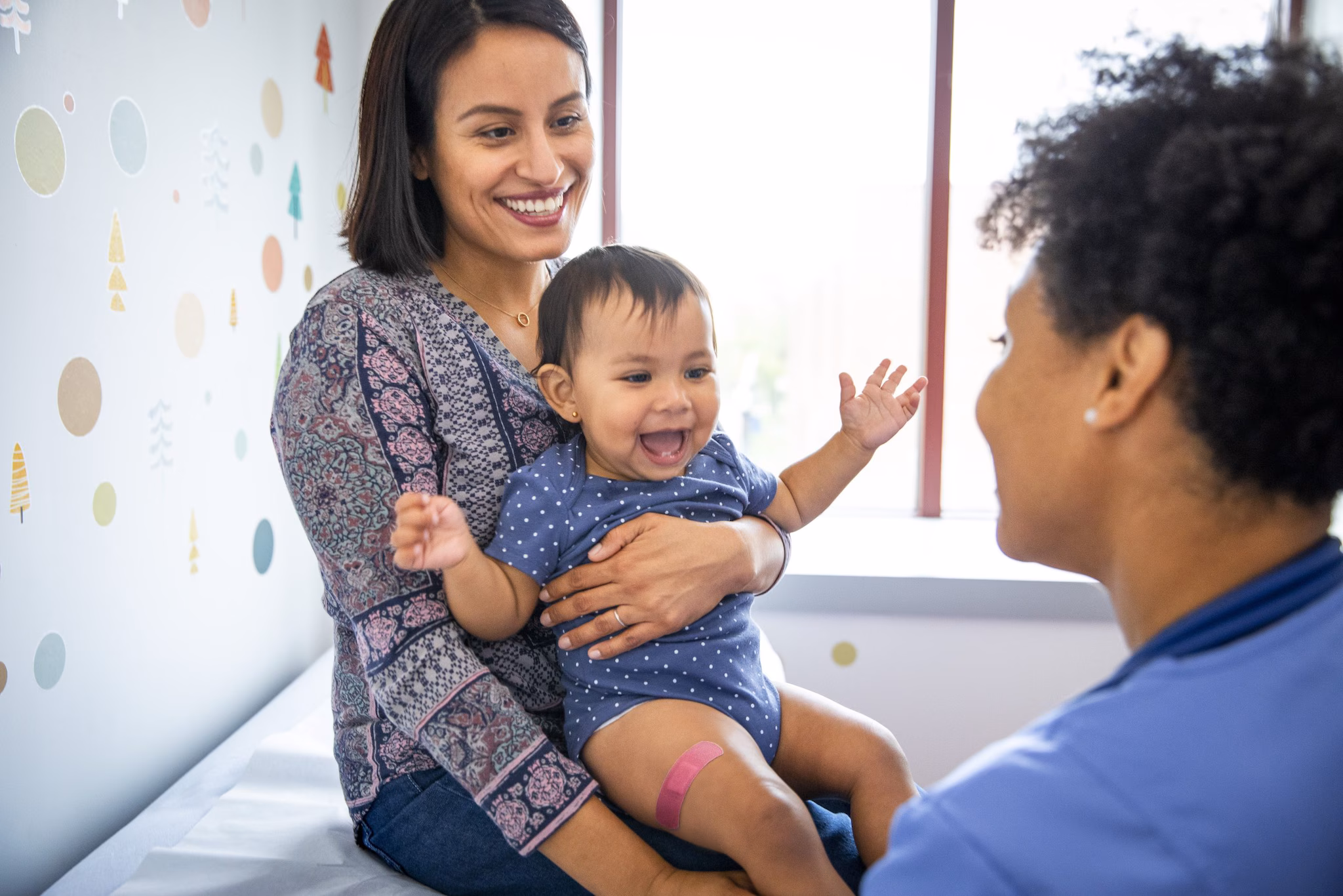 Parent and infant in Medical Office with Healthcare Provider Parent holding infant with a band aid on leg in medical office with healthcare provider post vaccination.