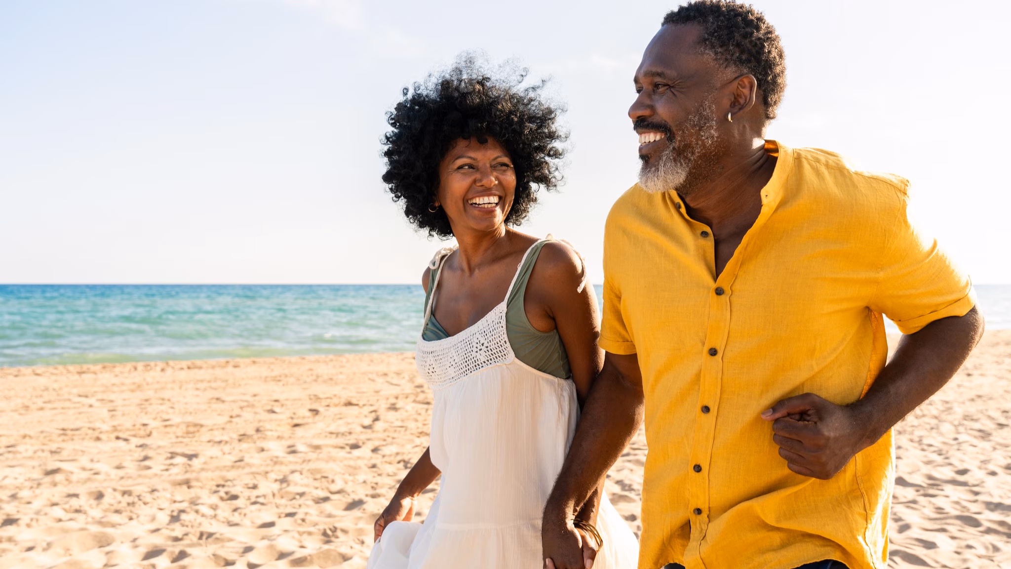 An older couple holding hands on the beach, walking on the sand.